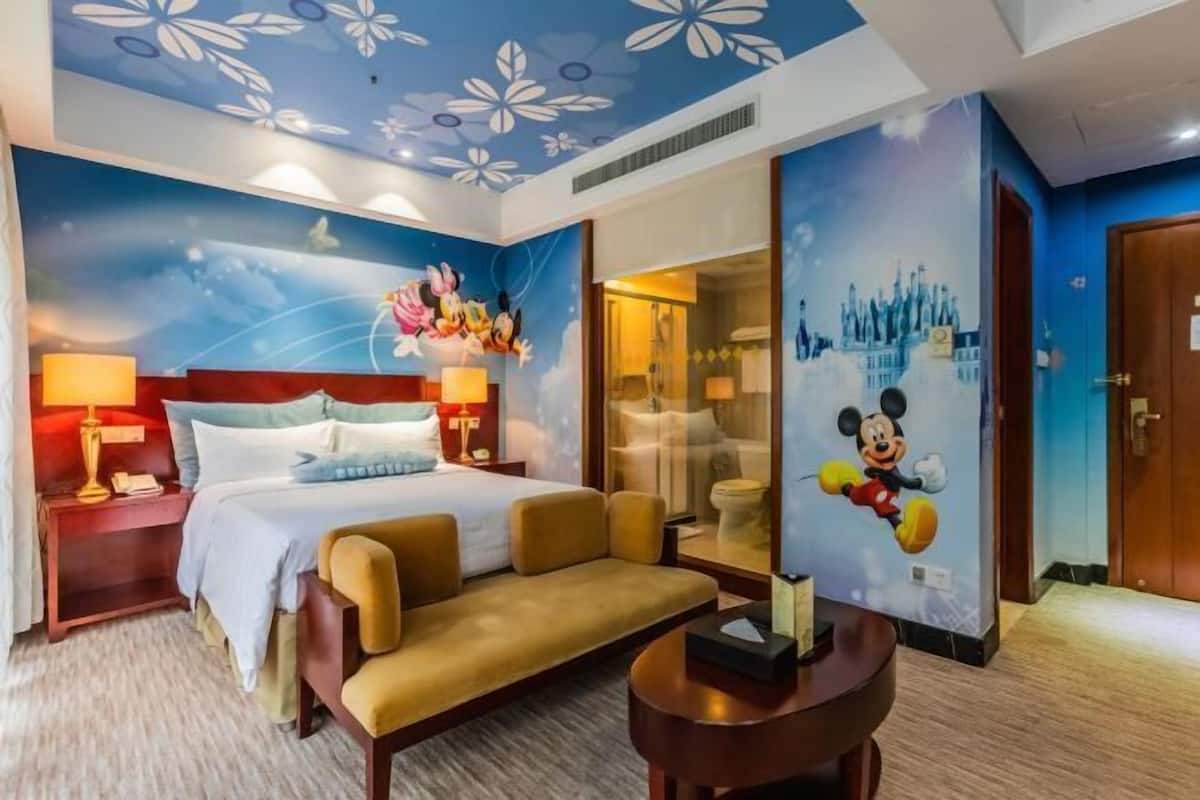 A hotel room with a bed, a sofa, a nightstand, a lamp, a mirror, and a Mickey Mouse mural.