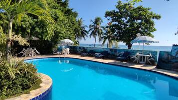 Piscina com espreguiçadeiras e guarda-sóis, cercada por vegetação tropical e um mural com uma cena de praia.