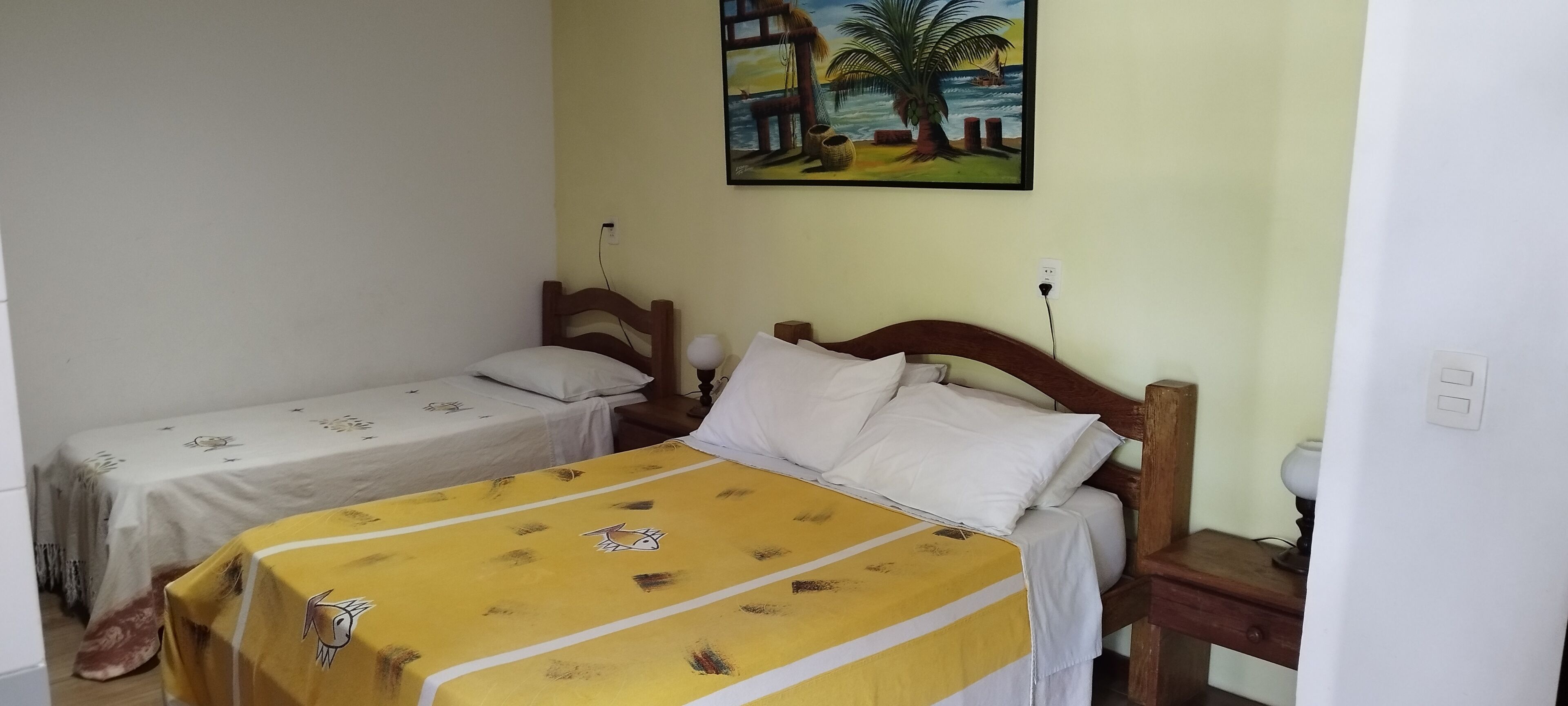 Ein Zimmer mit zwei Einzelbetten, eines mit gelber Bettdecke und das andere mit weißer Bettdecke. Über den Betten hängt ein Bild einer Strandansicht.