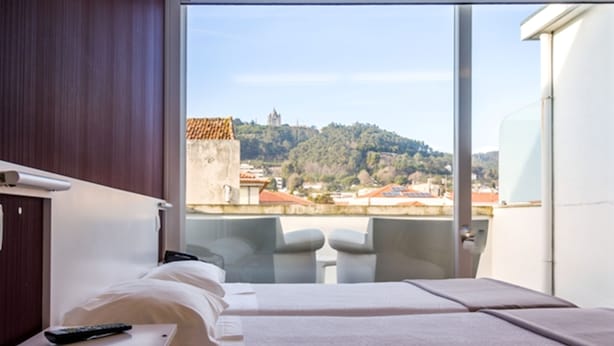 A hotel room with a bed, a small table with a remote control, and a view of a cityscape with buildings and a hill.