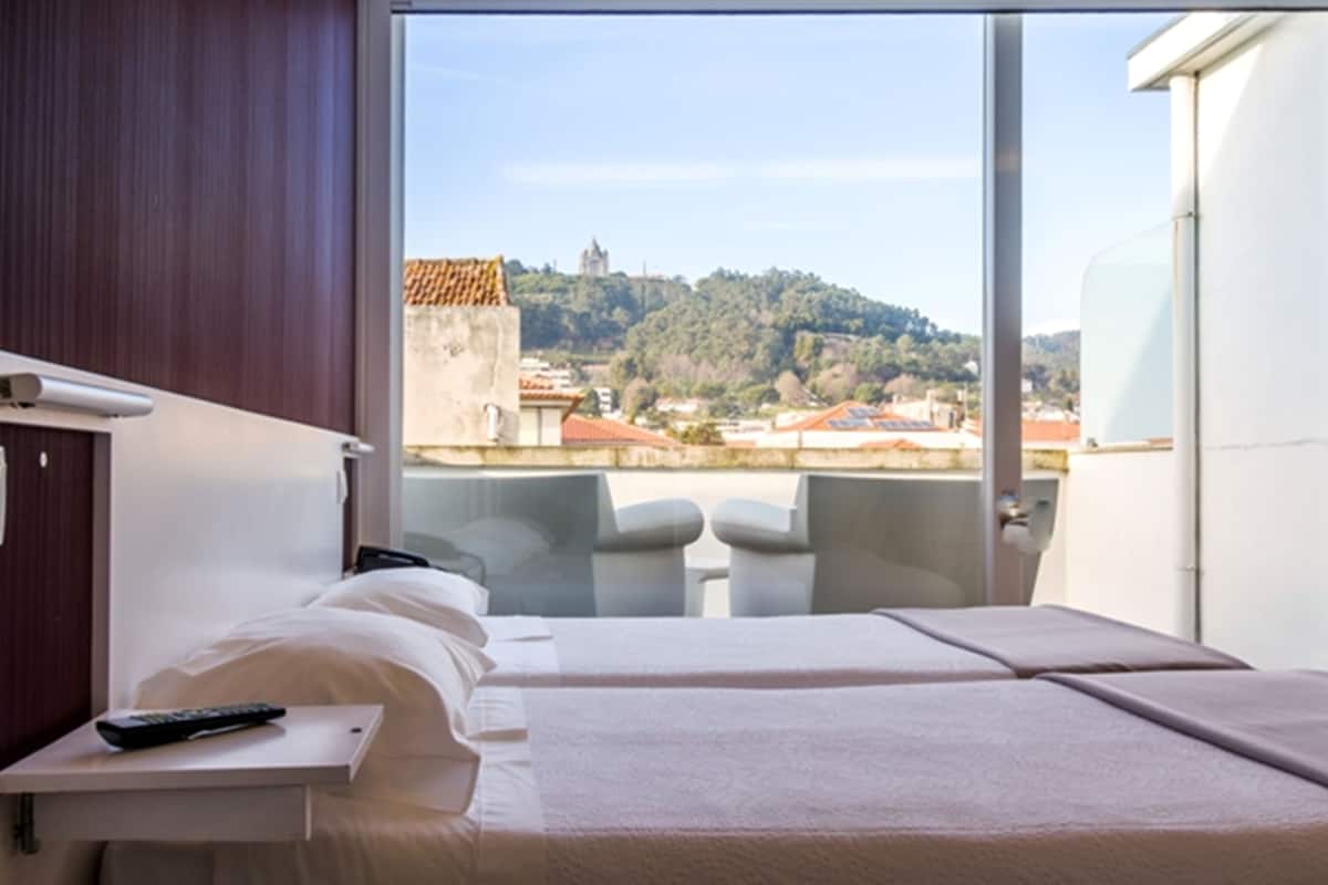 A hotel room with a bed, a small table with a remote control, and a view of a cityscape with buildings and a hill.
