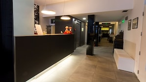 A hotel reception area with a dark reception desk, a waiting area with a bench, and a hallway leading to other rooms.