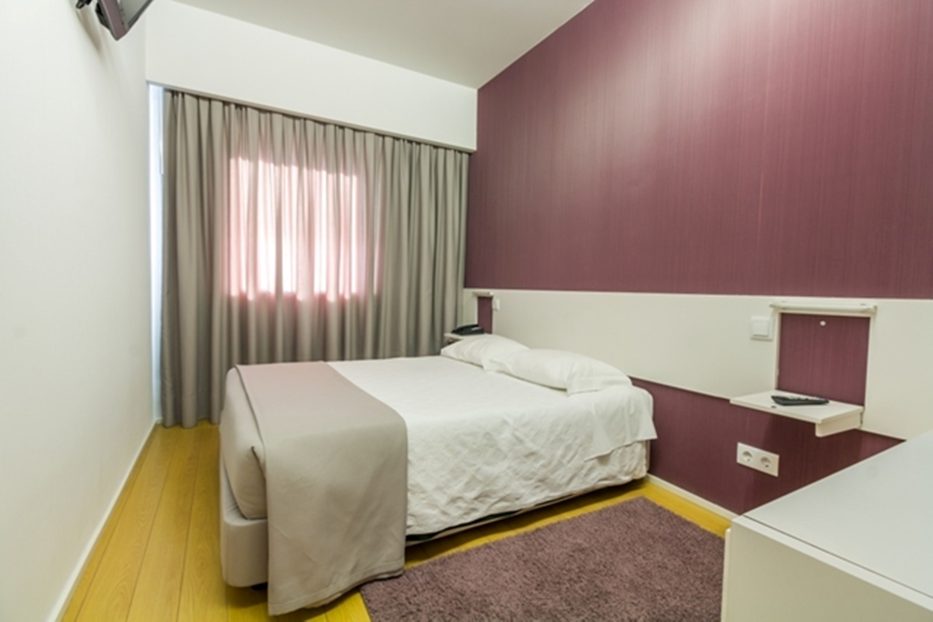 A hotel room with a bed, a bedside table, a nightstand, a window with curtains, and a wall-mounted shelf.