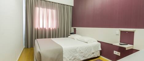 Una habitación de hotel con cama, mesita de noche, cortinas en la ventana y estante montado en la pared.