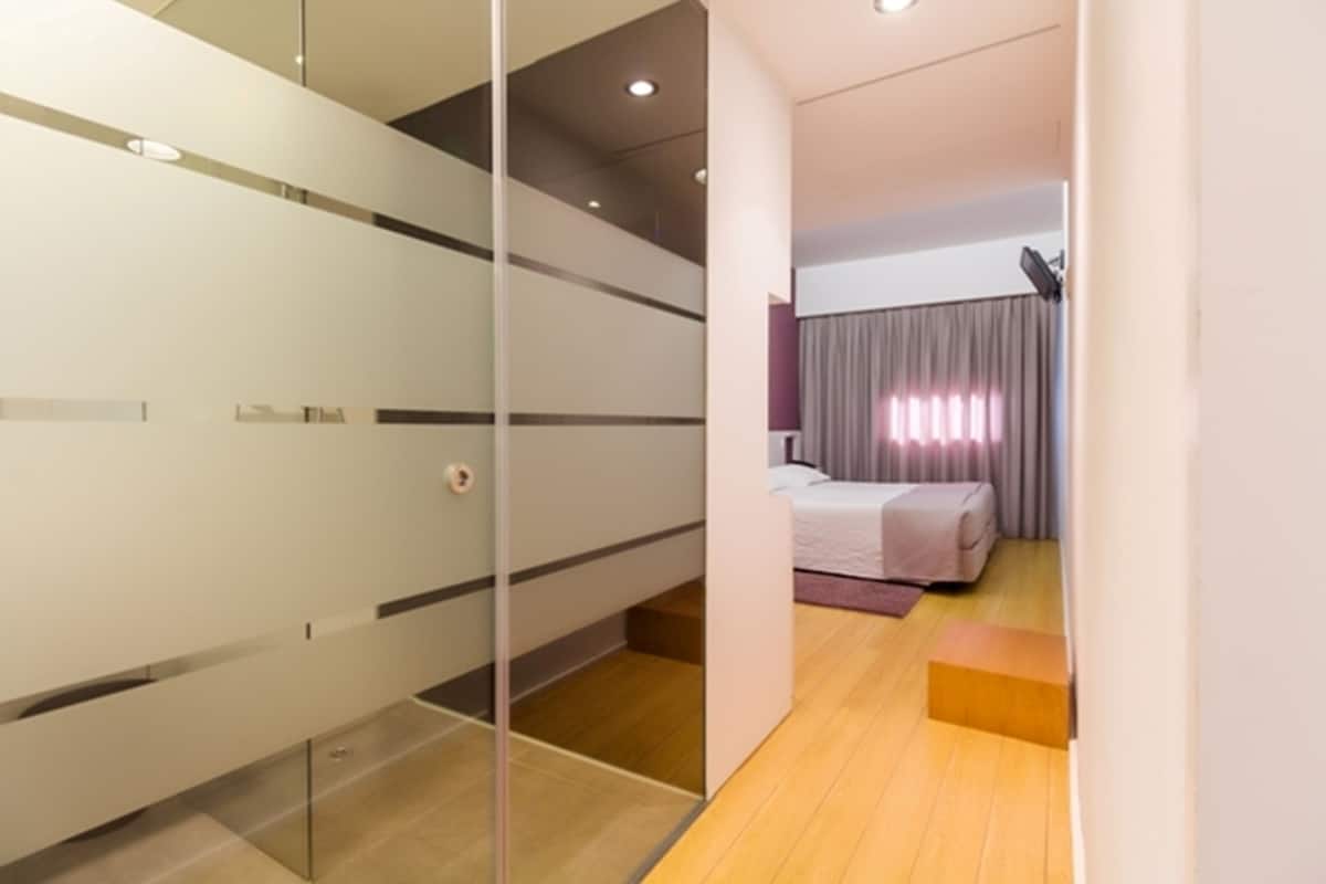 A modern hotel room with a glass partition, a bed with a white and purple bedspread, a wooden floor, and a small wooden bench.