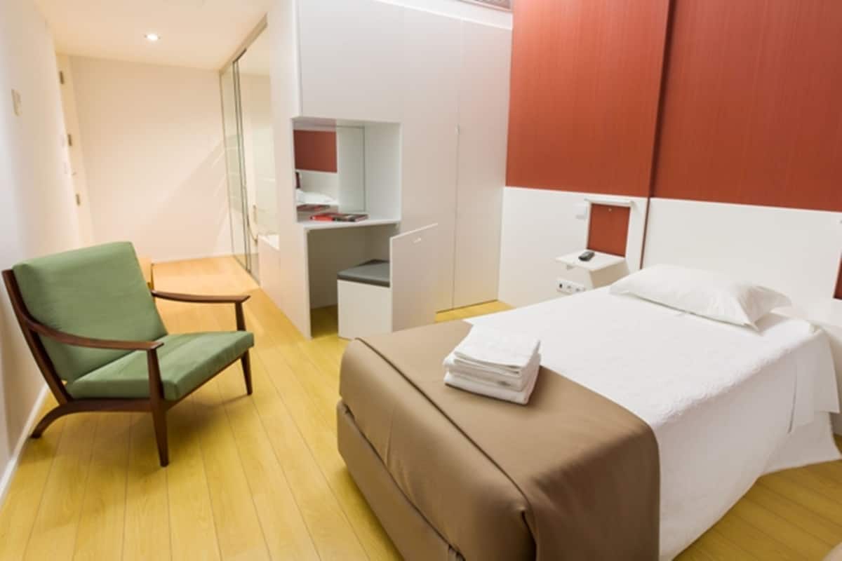 A hotel room with a bed, a green armchair, a small table, and a desk with a chair.