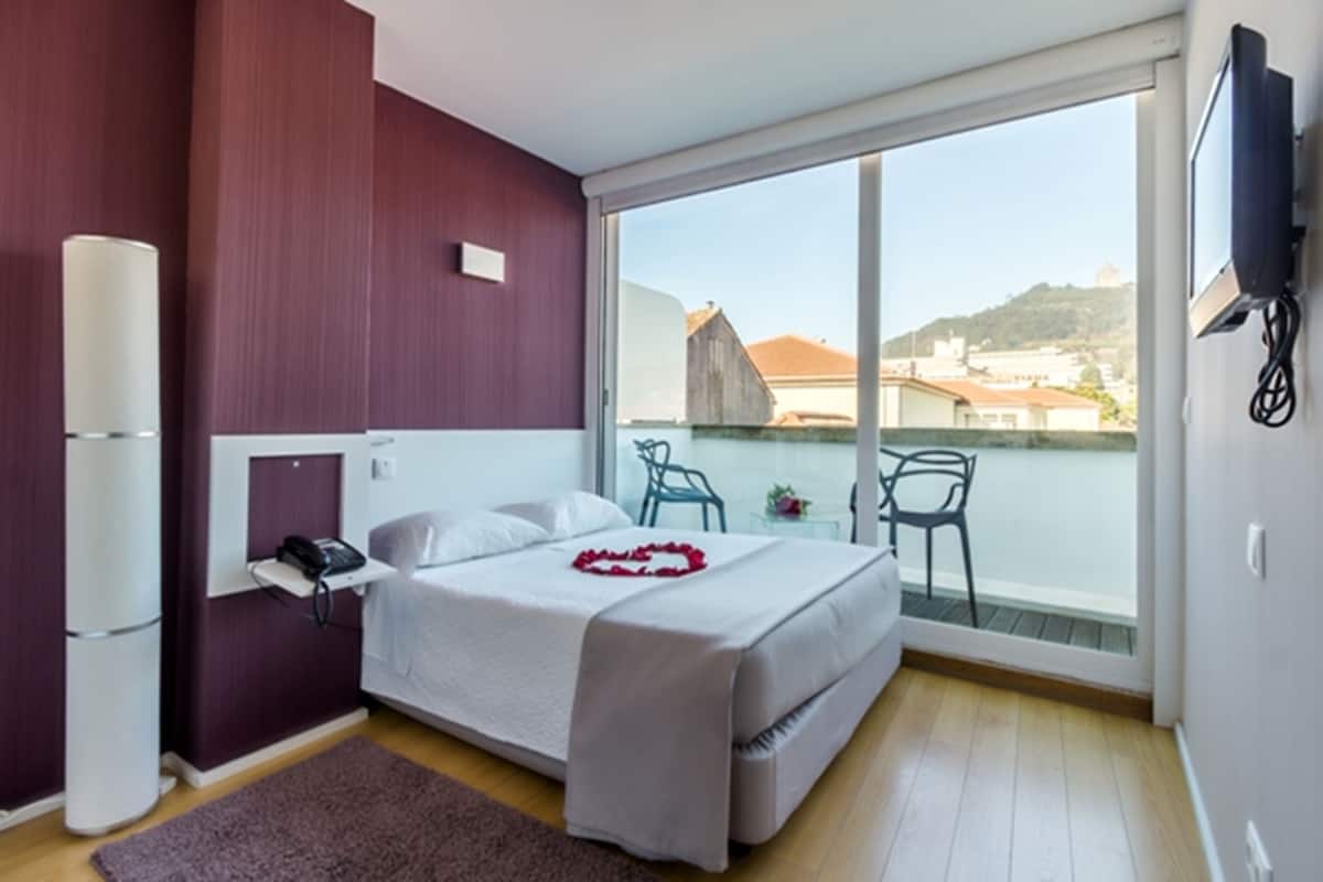 A hotel room with a bed, a bedside table with a phone, a chair by the window, and a TV mounted on the wall.