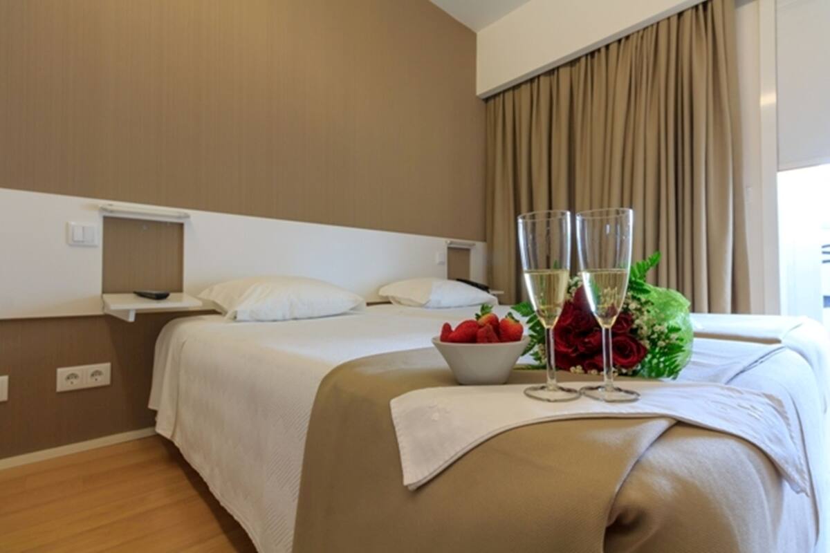 A hotel room with a bed, bedside tables, a nightstand, a window with curtains, a bouquet of flowers, and two glasses of champagne.