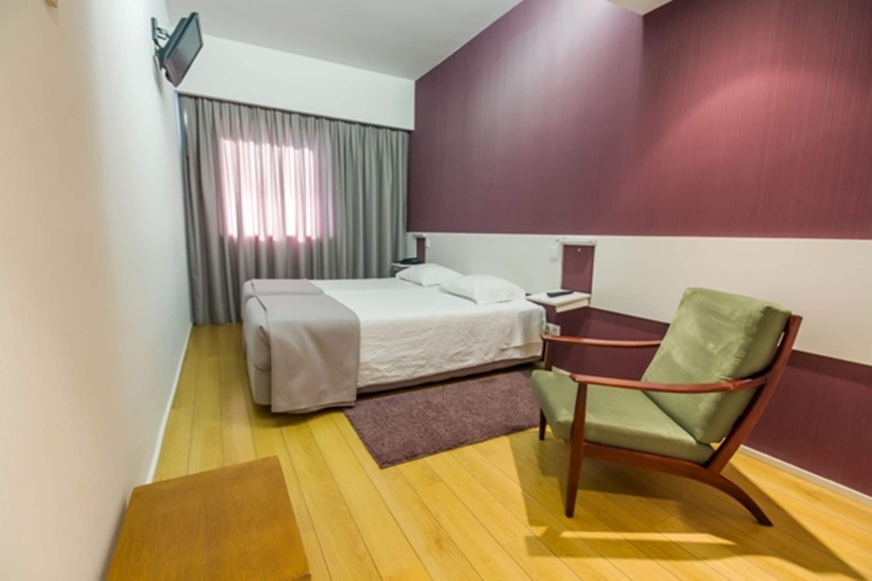 Una habitación de hotel con cama, una silla, escritorio, televisor y ventana con cortinas.