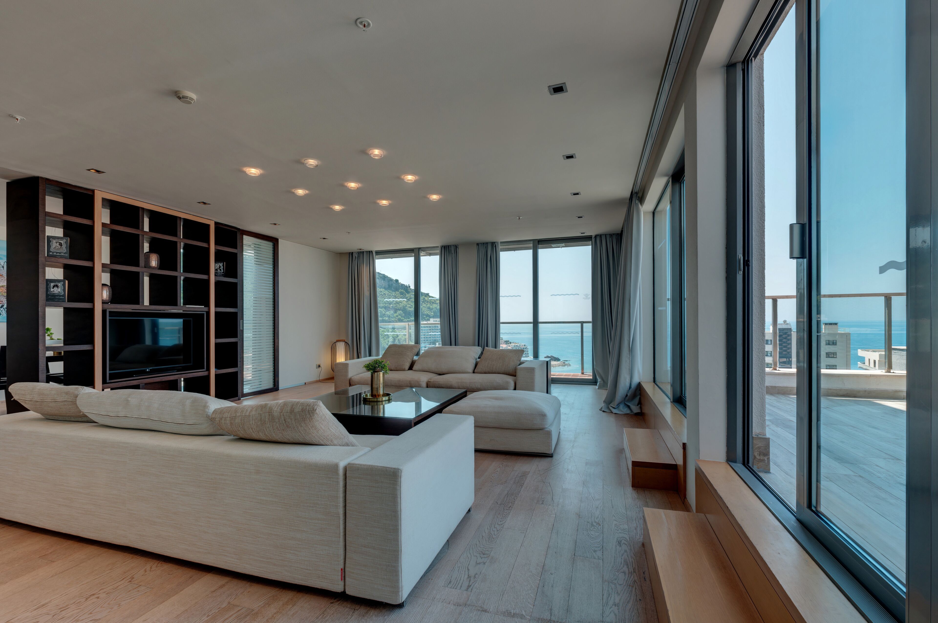 A modern living room with a large sofa, a coffee table, and a TV. There is a bookshelf and a large window with a view of the sea.