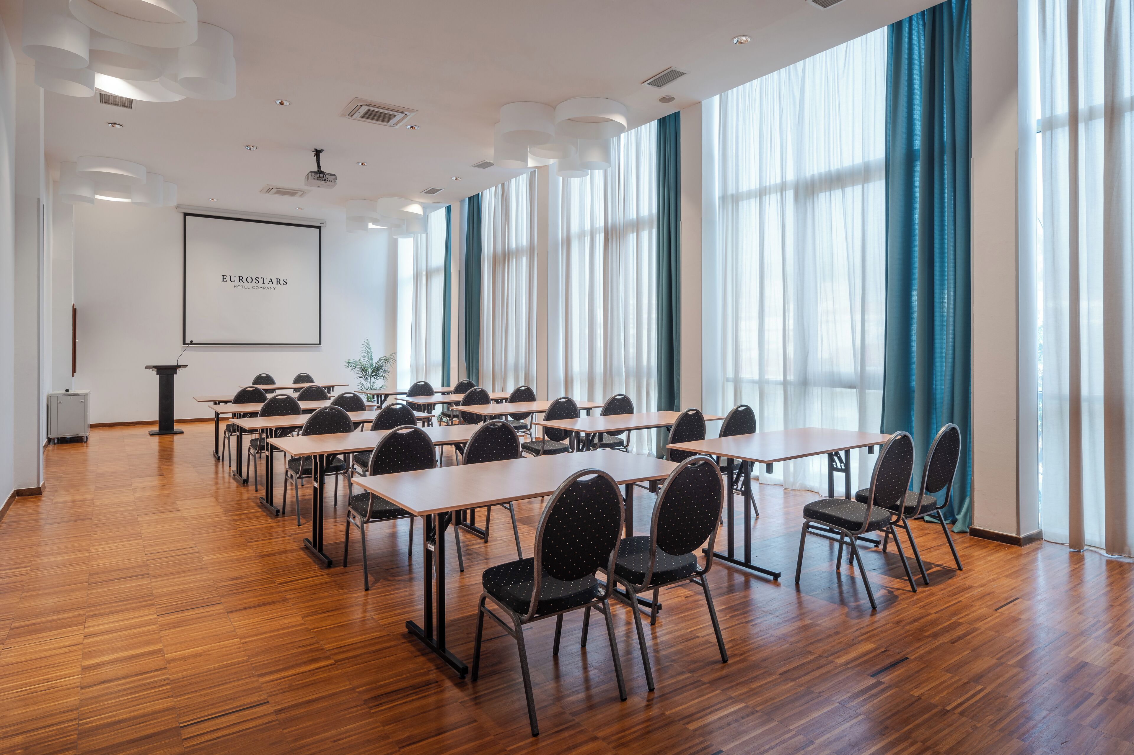 Una sala de conferències amb sòls de fusta, files de taules i cadires, grans finestres amb cortines blaves i una pantalla projectant "EUROSTARS HOTEL CORINTHIA".