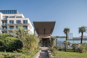 Edificio moderno con techo plano y fachada de vidrio. Escalones conducen a la entrada, rodeados de vegetación y palmeras.