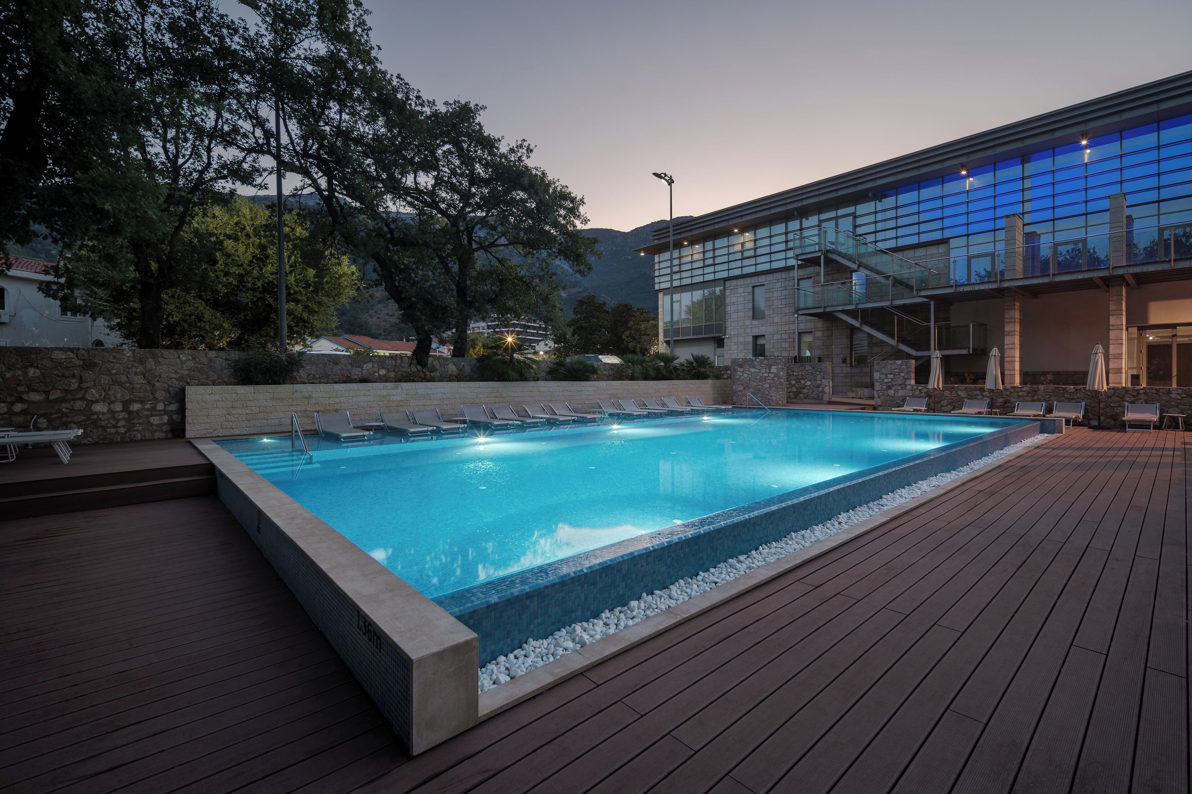 A rectangular swimming pool with a wooden deck, surrounded by lounge chairs and a stone wall.