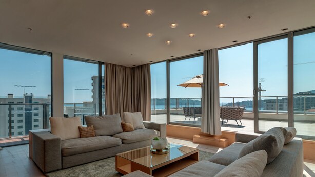 A modern living room with a sofa, a glass coffee table, and a view of the cityscape through large windows. - Eurostars Queen of Montenegro (Becici)