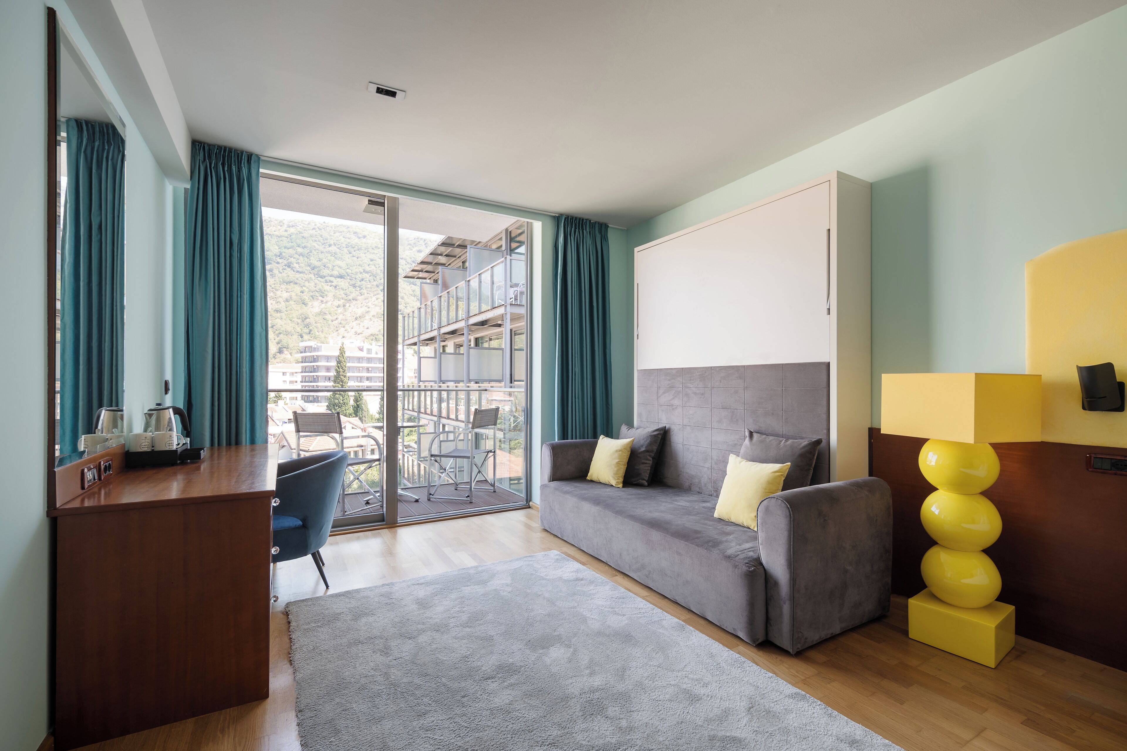 A modern hotel room with a grey sofa, a wooden desk, and a large window with curtains.