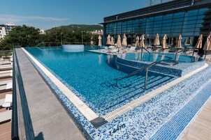 Una piscina infinita en la azotea, con sillones y sombrillas, rodeada por un edificio moderno con un borde de azulejos.