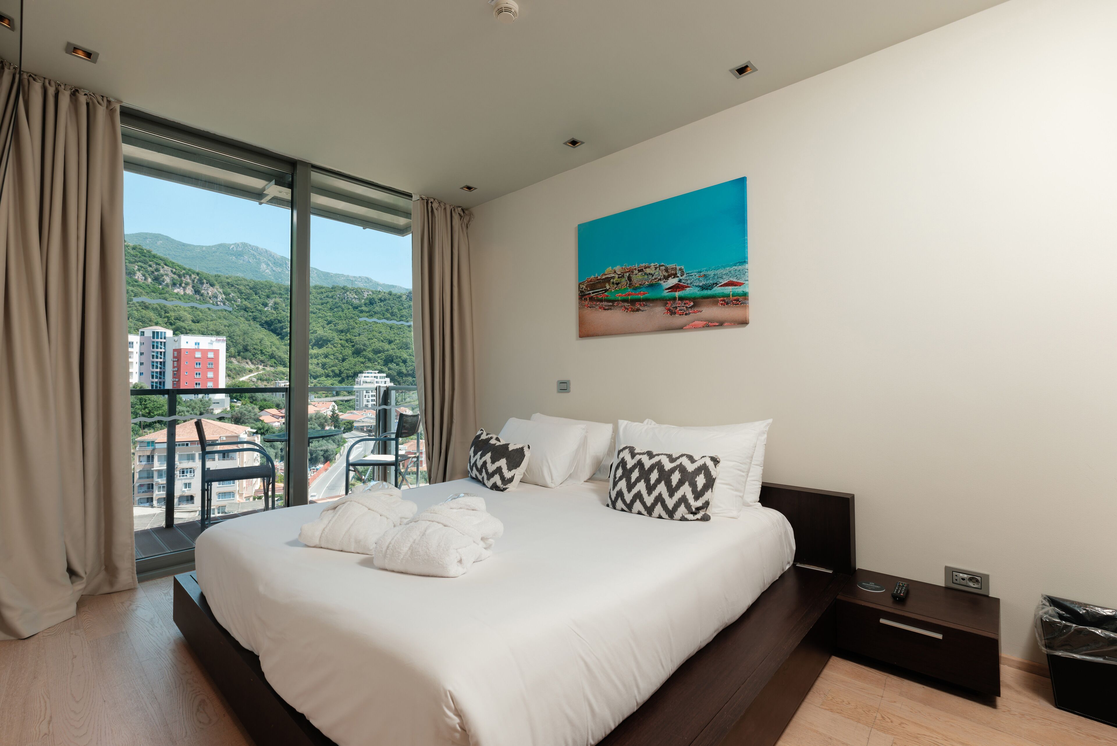 Ein Hotelzimmer mit einem großen Bett, einem Strandbild und Blick auf Gebäude und Berge durch das Fenster.