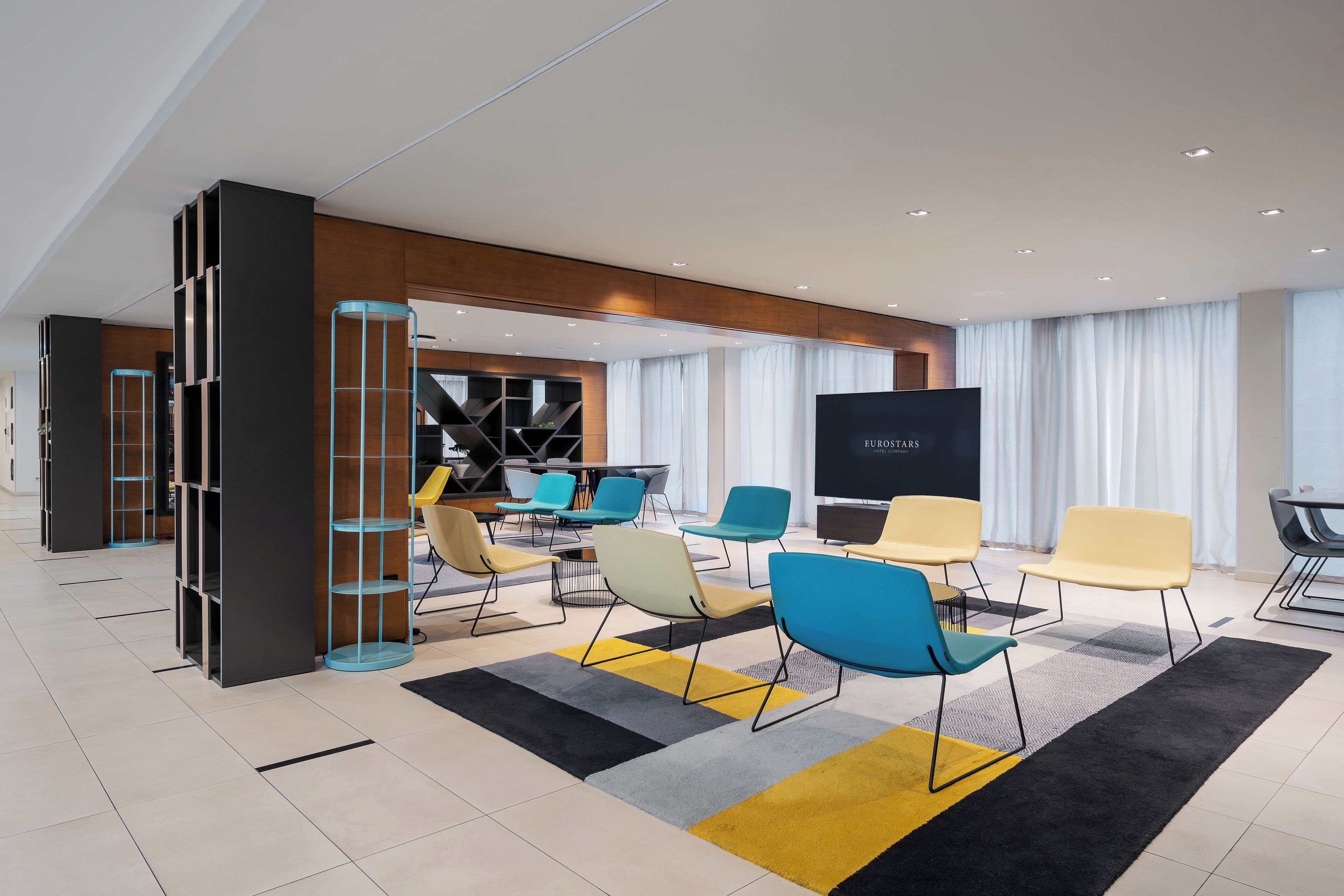 A modern hotel lobby with a large TV screen, multiple seating areas with colorful chairs, and a large window with curtains.