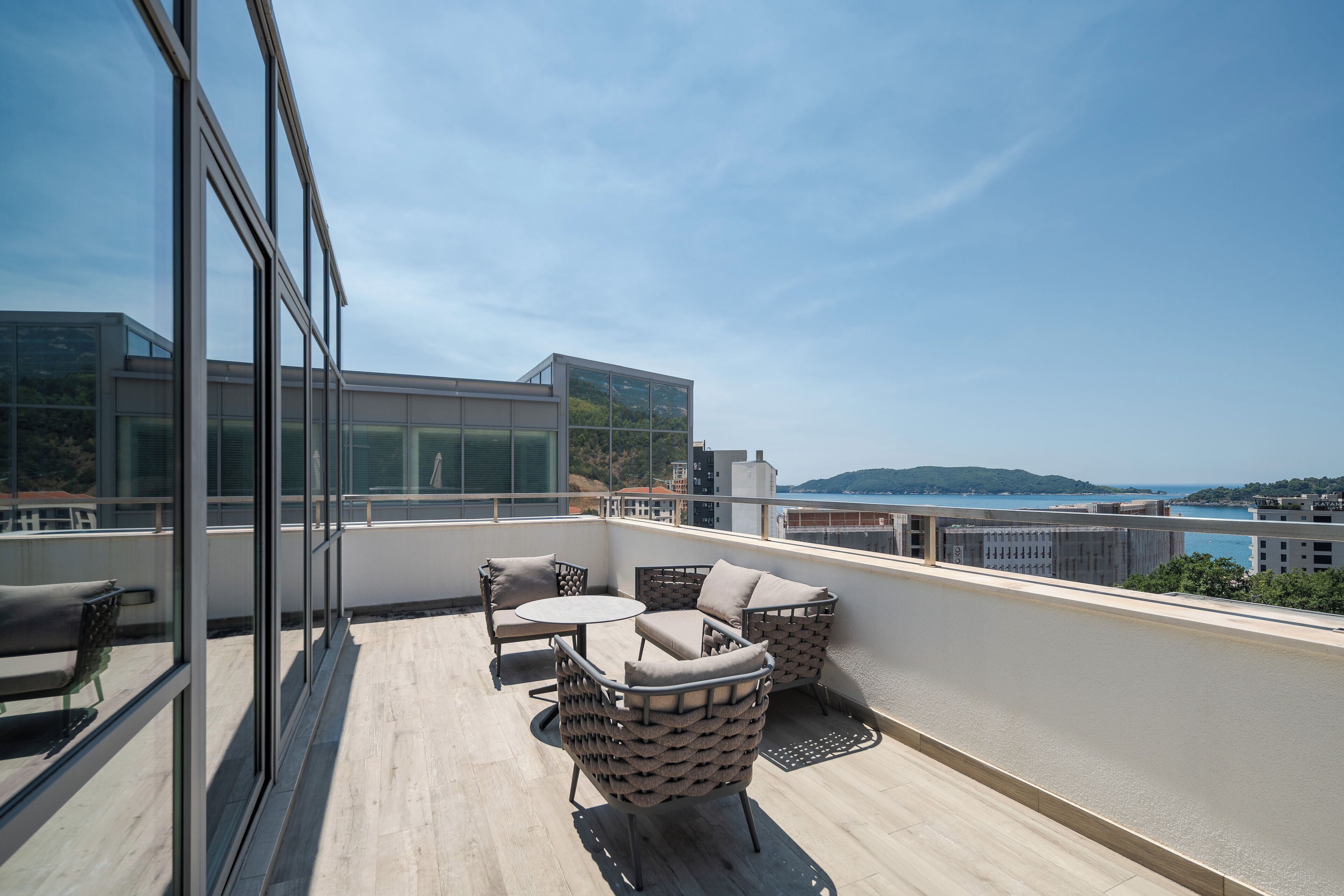 A rooftop terrace with outdoor seating, a view of the sea, and modern buildings in the background.