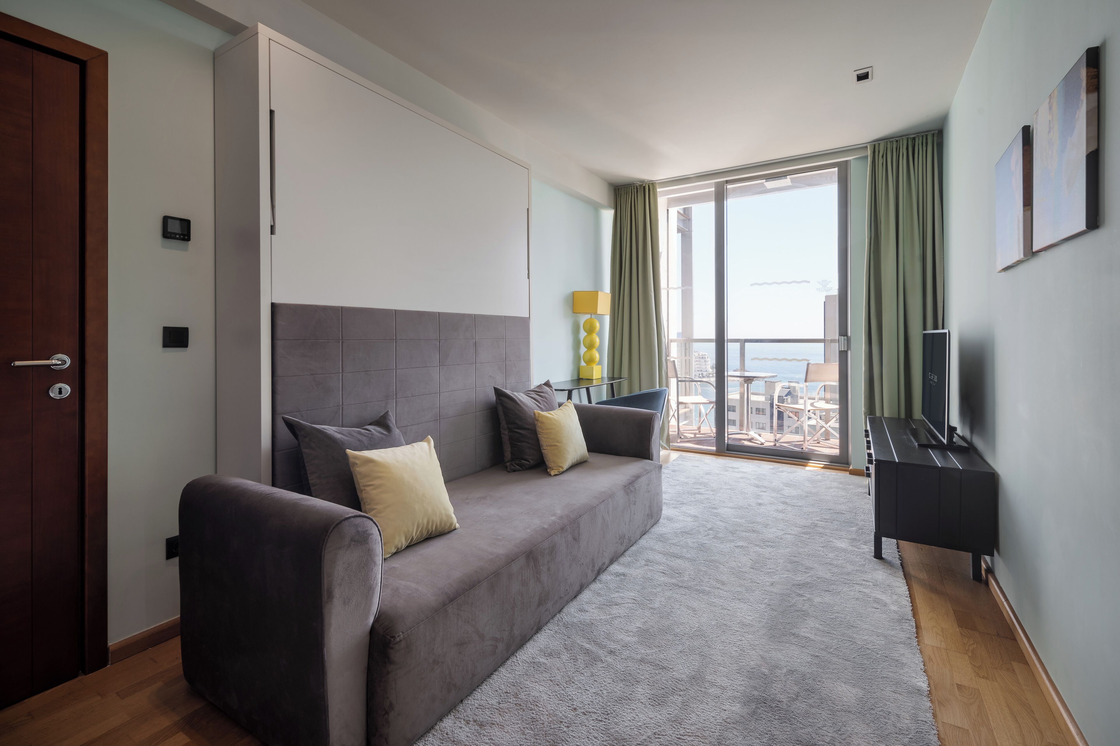 A modern living room with a grey sofa, a flat-screen TV, and a balcony with a view.