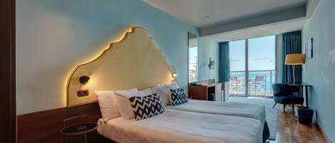 A hotel room with two beds, a desk, a chair, and a view of the sea.