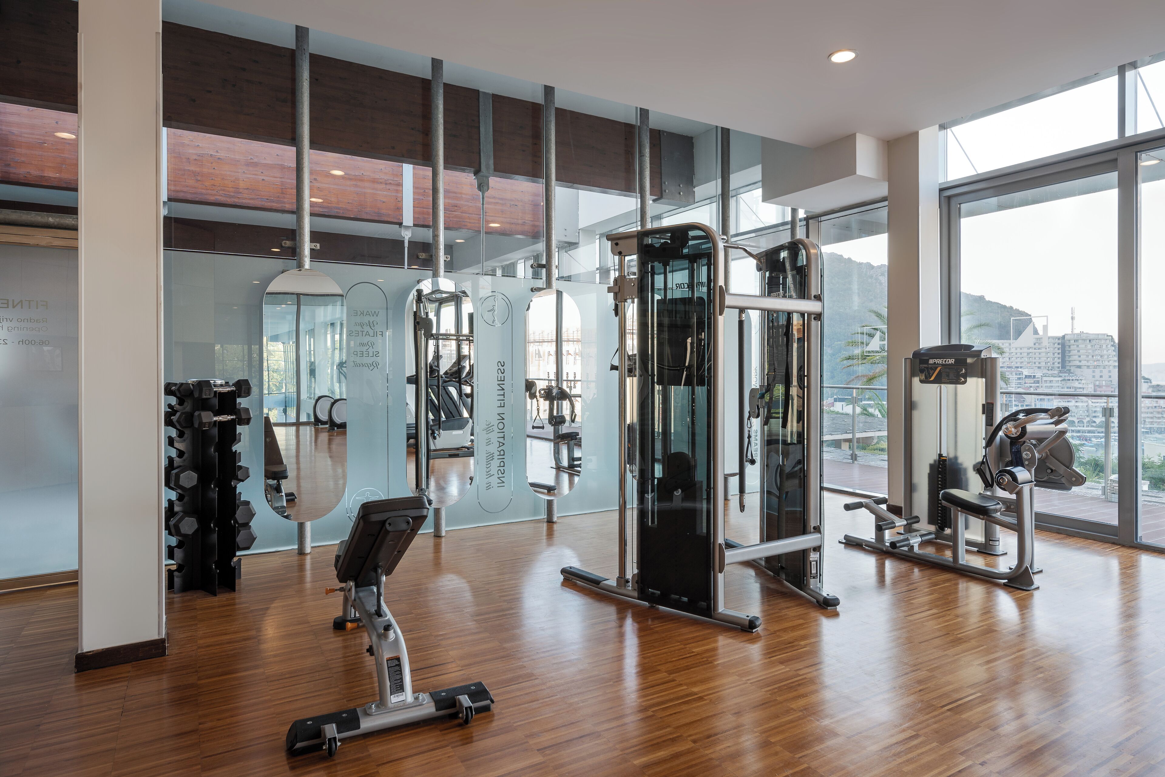 A modern gym with various exercise equipment, large windows, and a wooden floor.