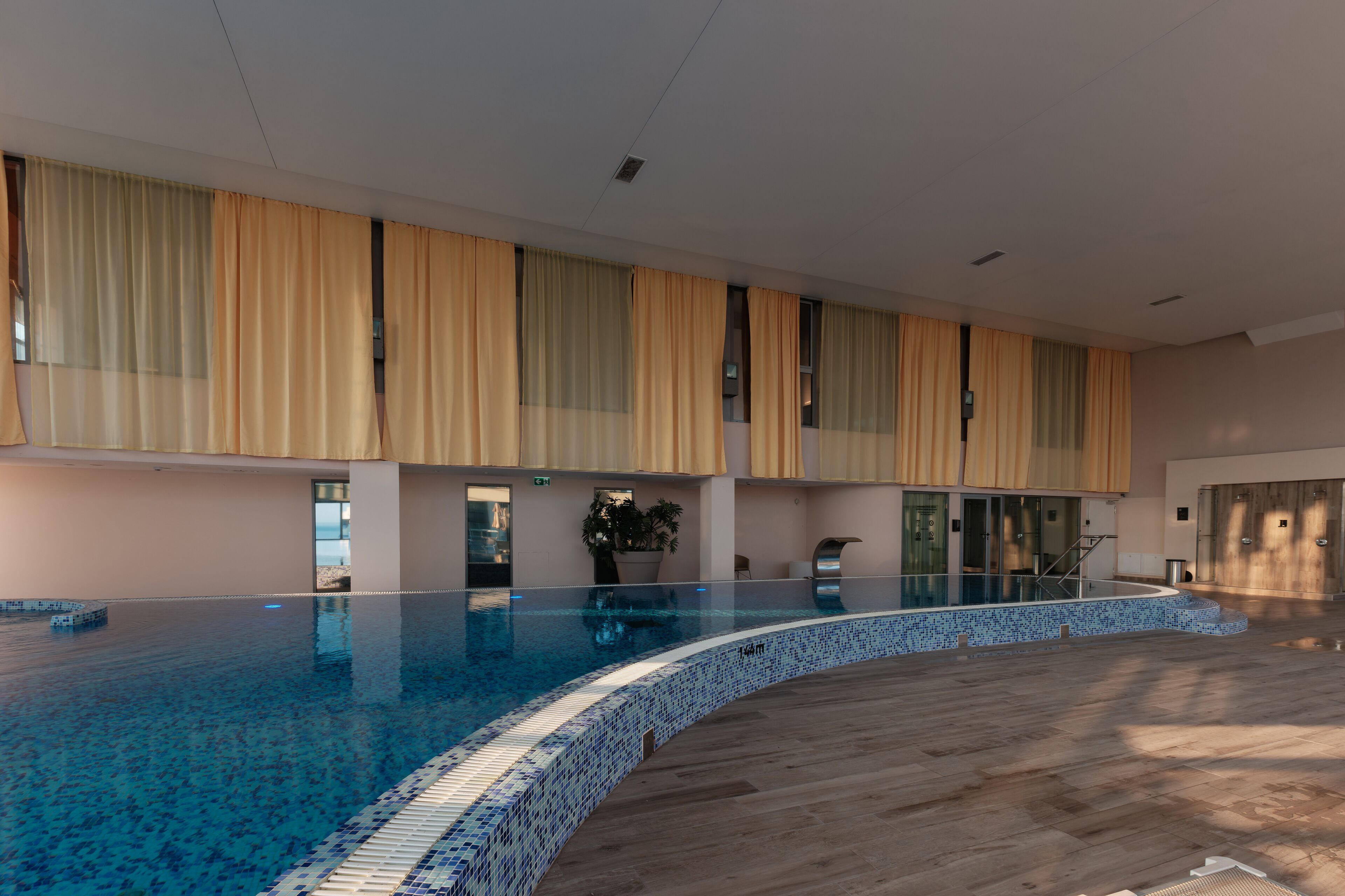A large indoor swimming pool with a tiled edge, surrounded by a wooden deck and beige curtains.