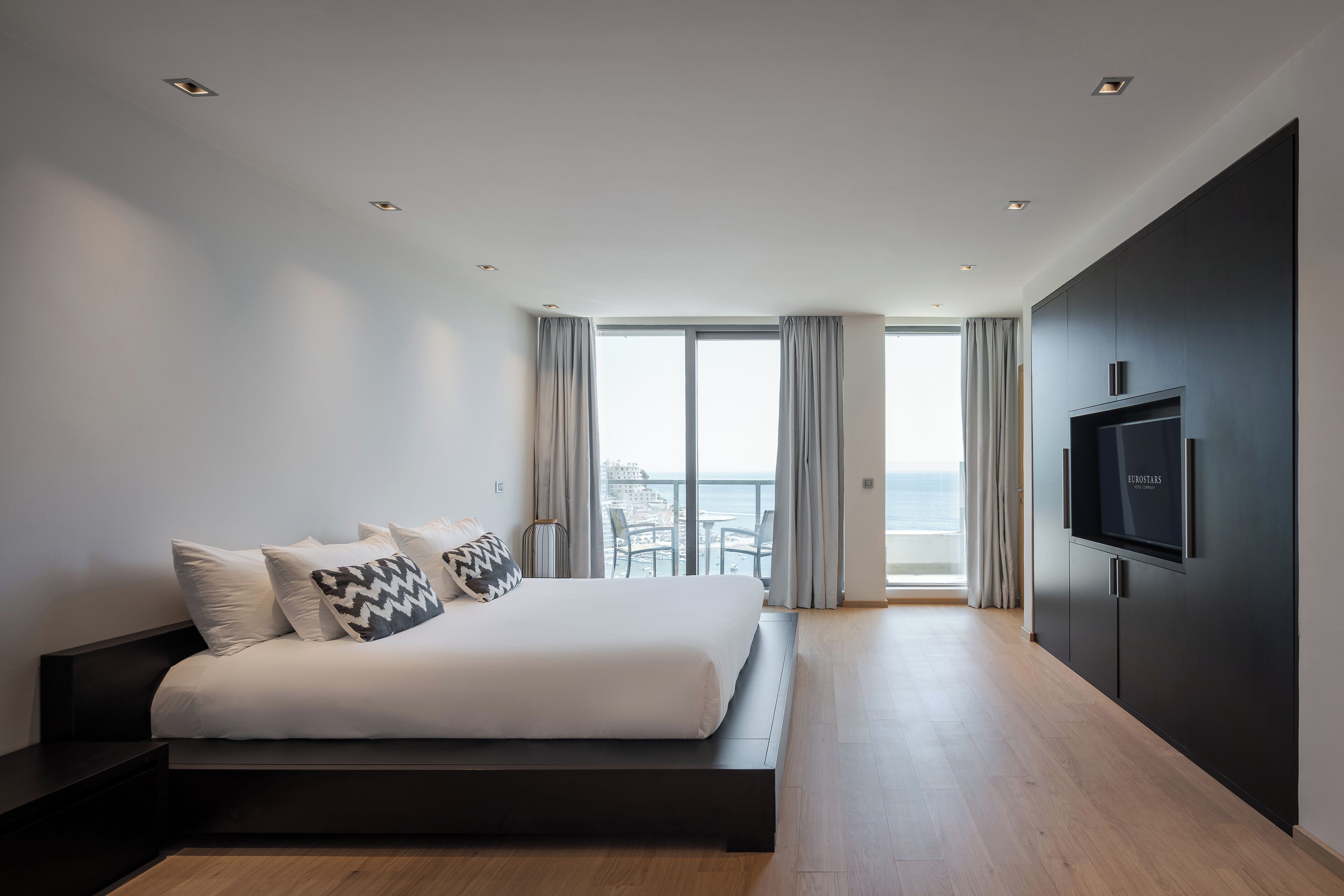 A modern bedroom with a large bed, a TV mounted on the wall, and a balcony with curtains.
