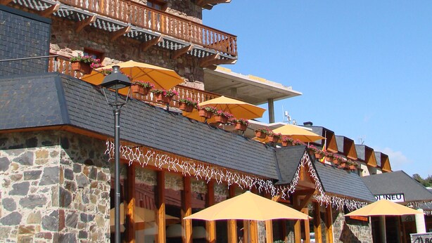 A building with a stone facade, outdoor seating with umbrellas, and a balcony with wooden railings.