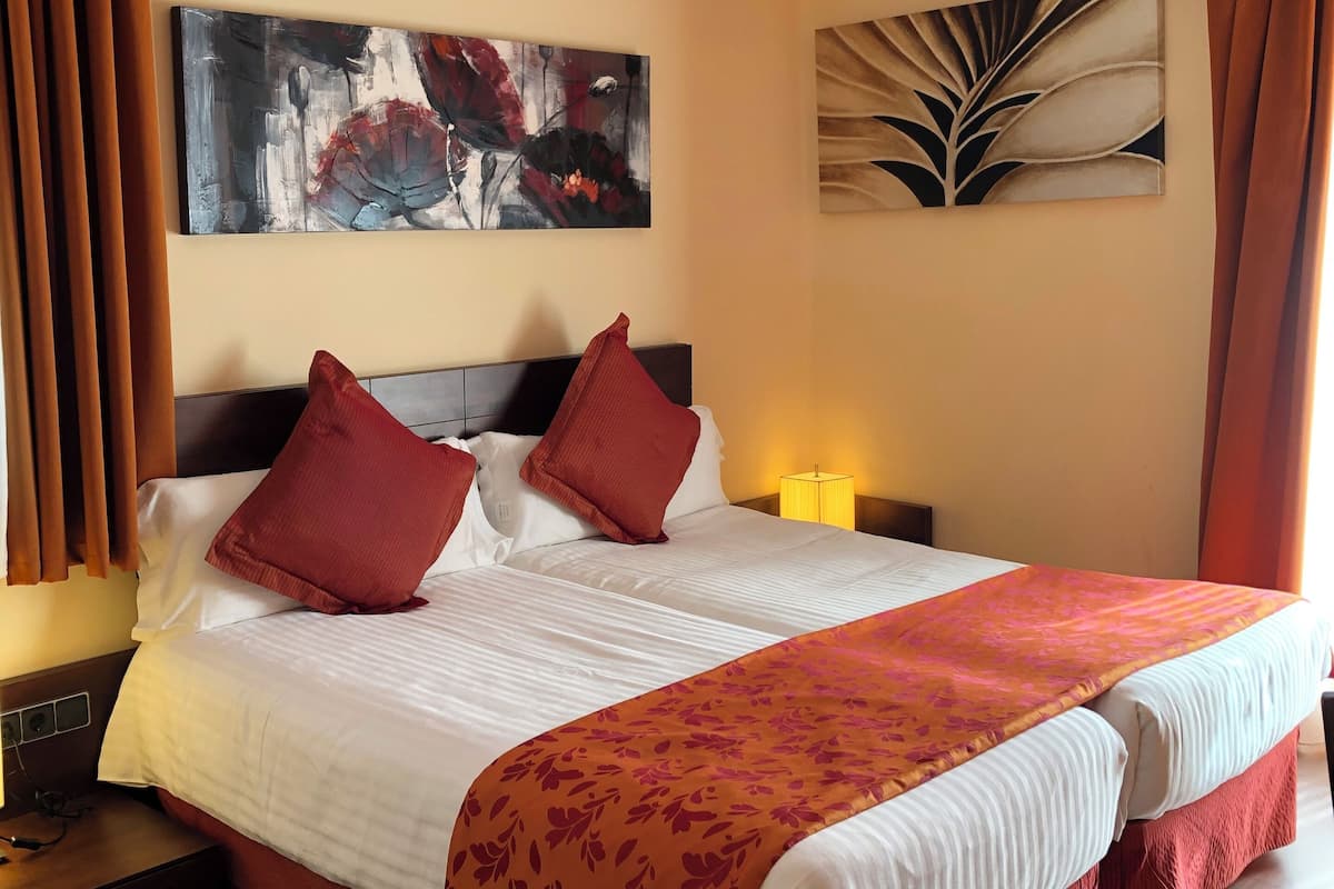 A hotel room with a large bed, two red pillows, a white comforter, and a floral throw blanket. There are two wall-mounted paintings, a nightstand with a lamp, and a wooden floor.