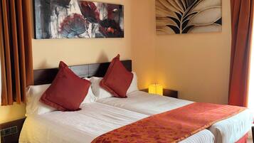 A hotel room with a large bed, two red pillows, a white comforter, and a floral throw blanket. There are two wall-mounted paintings, a nightstand with a lamp, and a wooden floor.