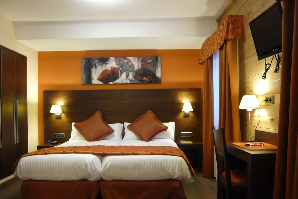 A hotel room with a double bed, two bedside tables with lamps, a TV mounted on the wall, and a desk with a chair.