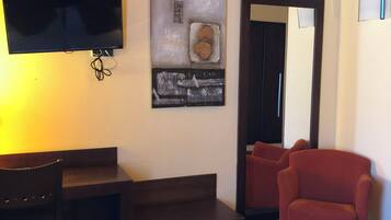 Quarto de hotel com cama, escrivaninha, cadeira, televisão e um quadro na parede.