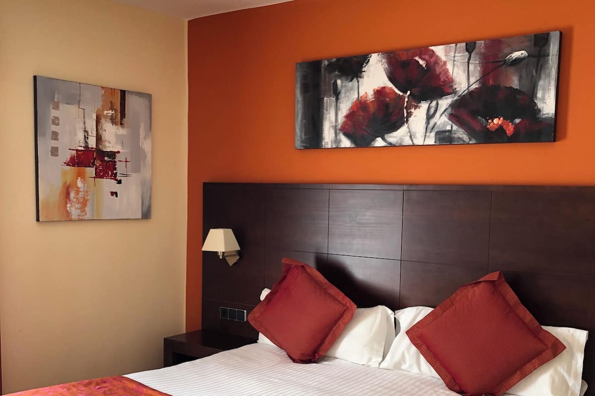 A hotel room with a large bed, two red pillows, a wooden headboard, a nightstand with a lamp, and two wall art pieces.
