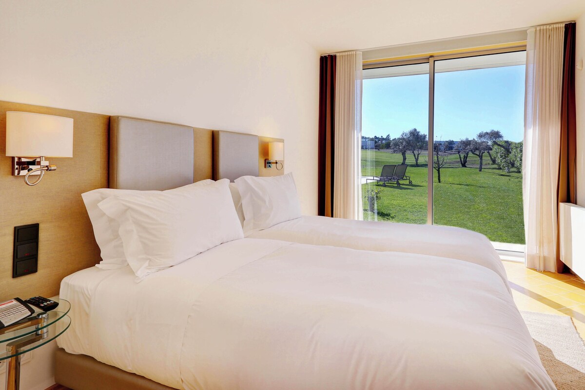 A hotel room with a large bed, a TV, a bedside table with a remote, and a window with a view of a grassy area.