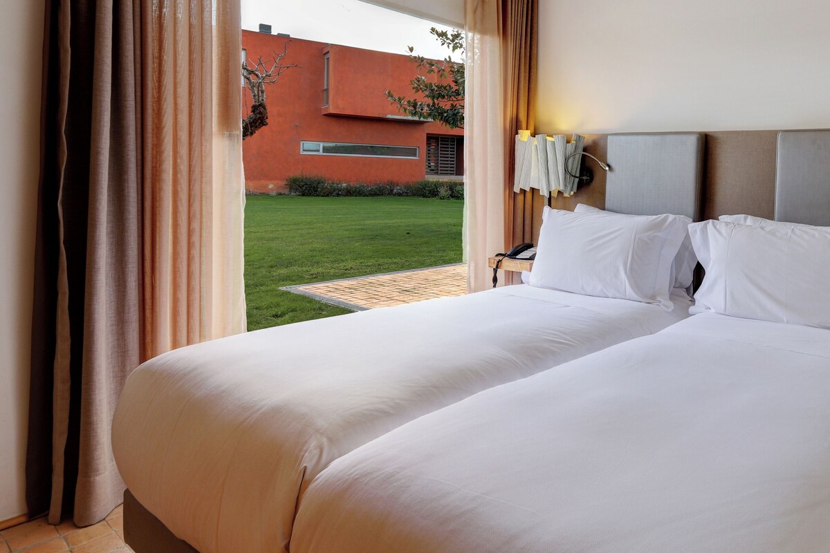 A double bed with white linens, a bedside table with a lamp, and a view of a building and lawn outside.