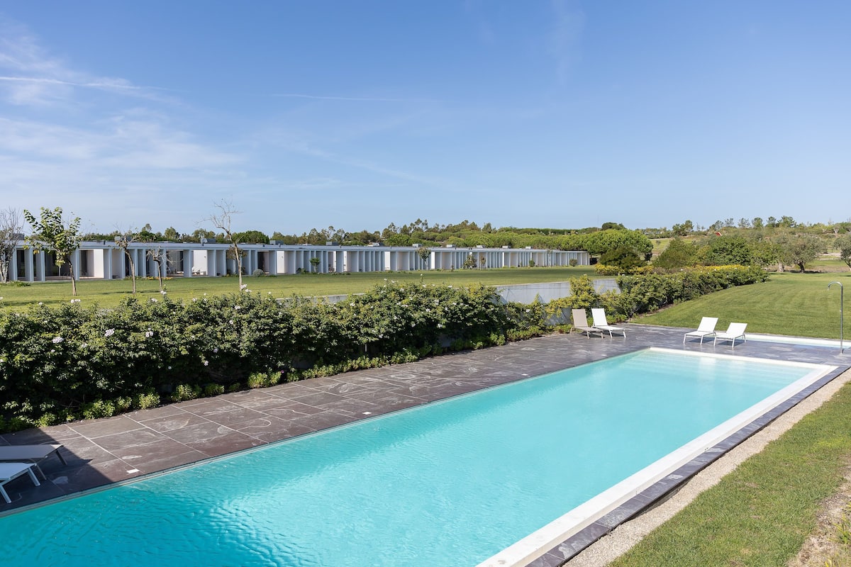 Une piscine rectangulaire bordée d’une terrasse carrelée, entourée d’une pelouse et avec, en arrière-plan, une rangée de bâtiments blancs.