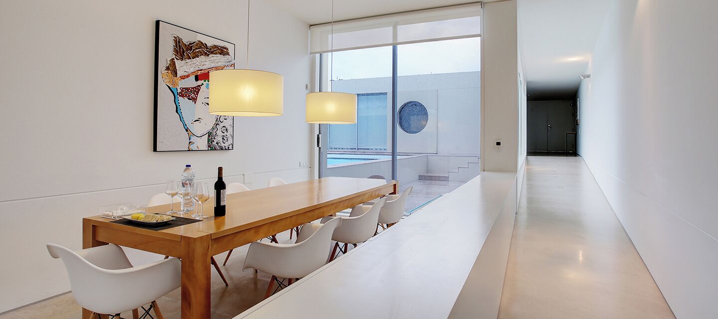 Un espace de repas moderne avec une longue table en bois, des chaises blanches et deux lampes suspendues. Une grande fenêtre offre une vue sur une piscine et un escalier menant à un niveau supérieur.