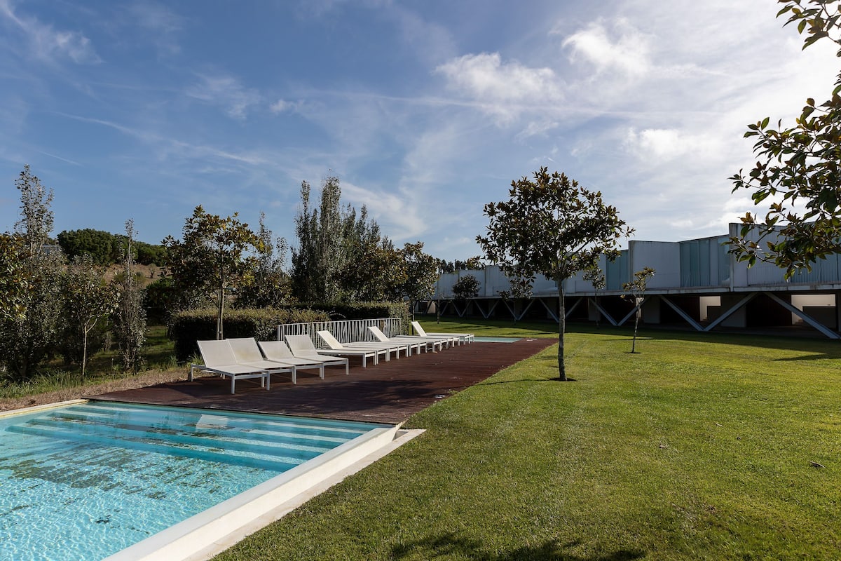 Une piscine avec des chaises longues, une pelouse et un bâtiment en arrière-plan.
