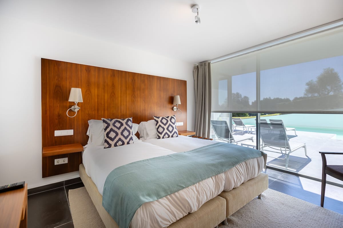 Une chambre d’hôtel moderne dotée d’un grand lit, d’une tête de lit en bois, de deux lampes de chevet, d’une vue sur l’océan grâce à de grandes fenêtres et d’un balcon avec des meubles d’extérieur.