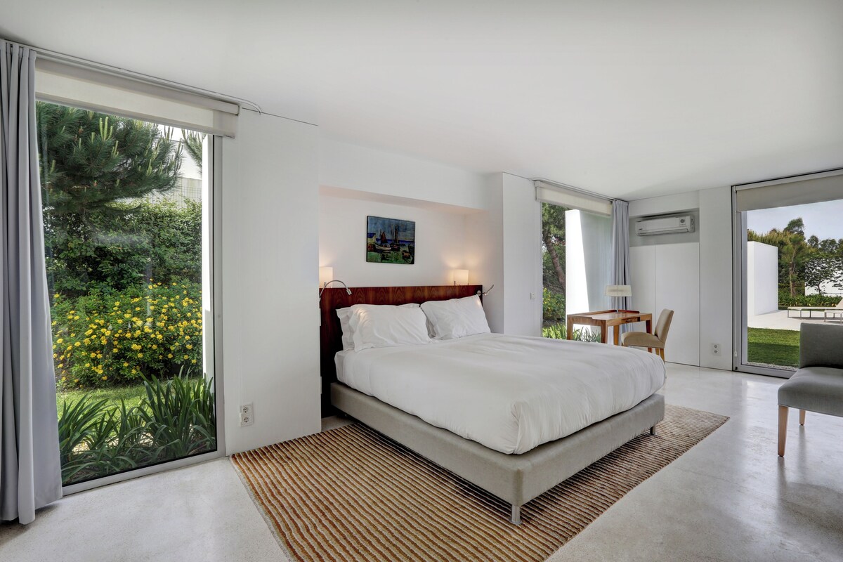 A modern bedroom with a large bed, a desk, and a view of greenery.