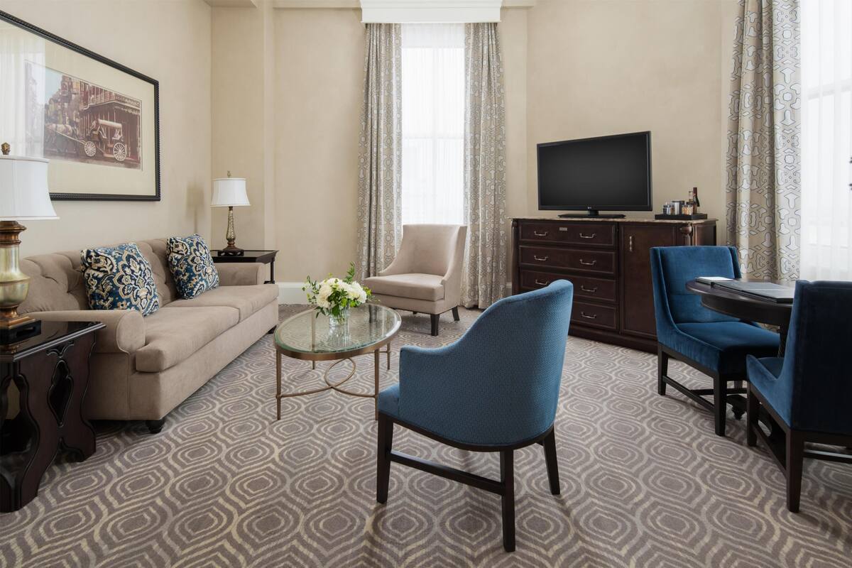 Een hotelkamer met een bank, fauteuils, een televisie, een kleine tafel met een vaas en een kamerbreed tapijt met dessin.
