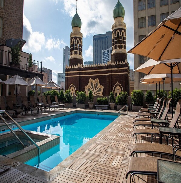 Zona de piscina con sillones de descanso y sombrillas, rodeada por un edificio con dos torres abovedadas.