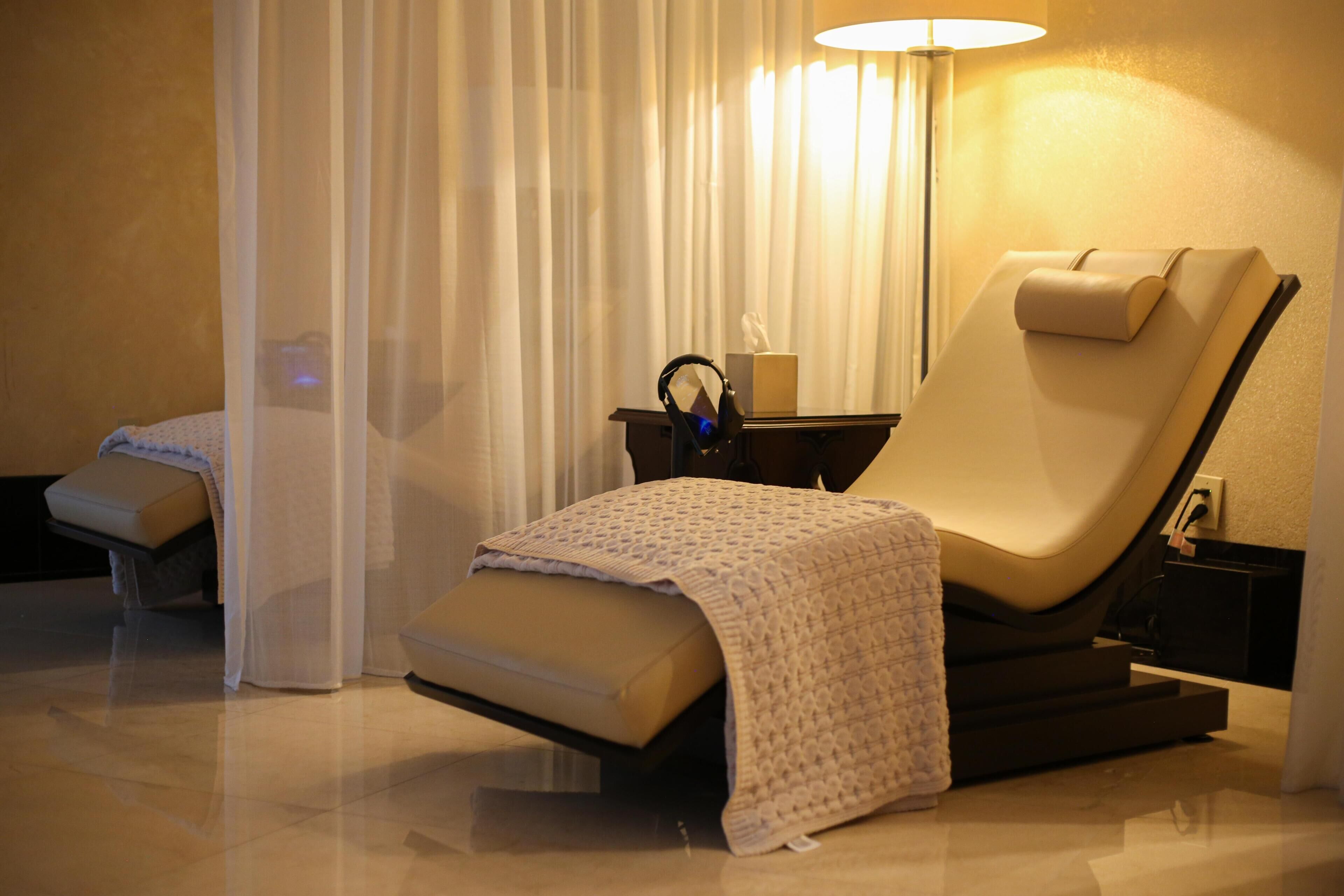A massage chair with a footrest, a small table with a lamp, and a curtain.