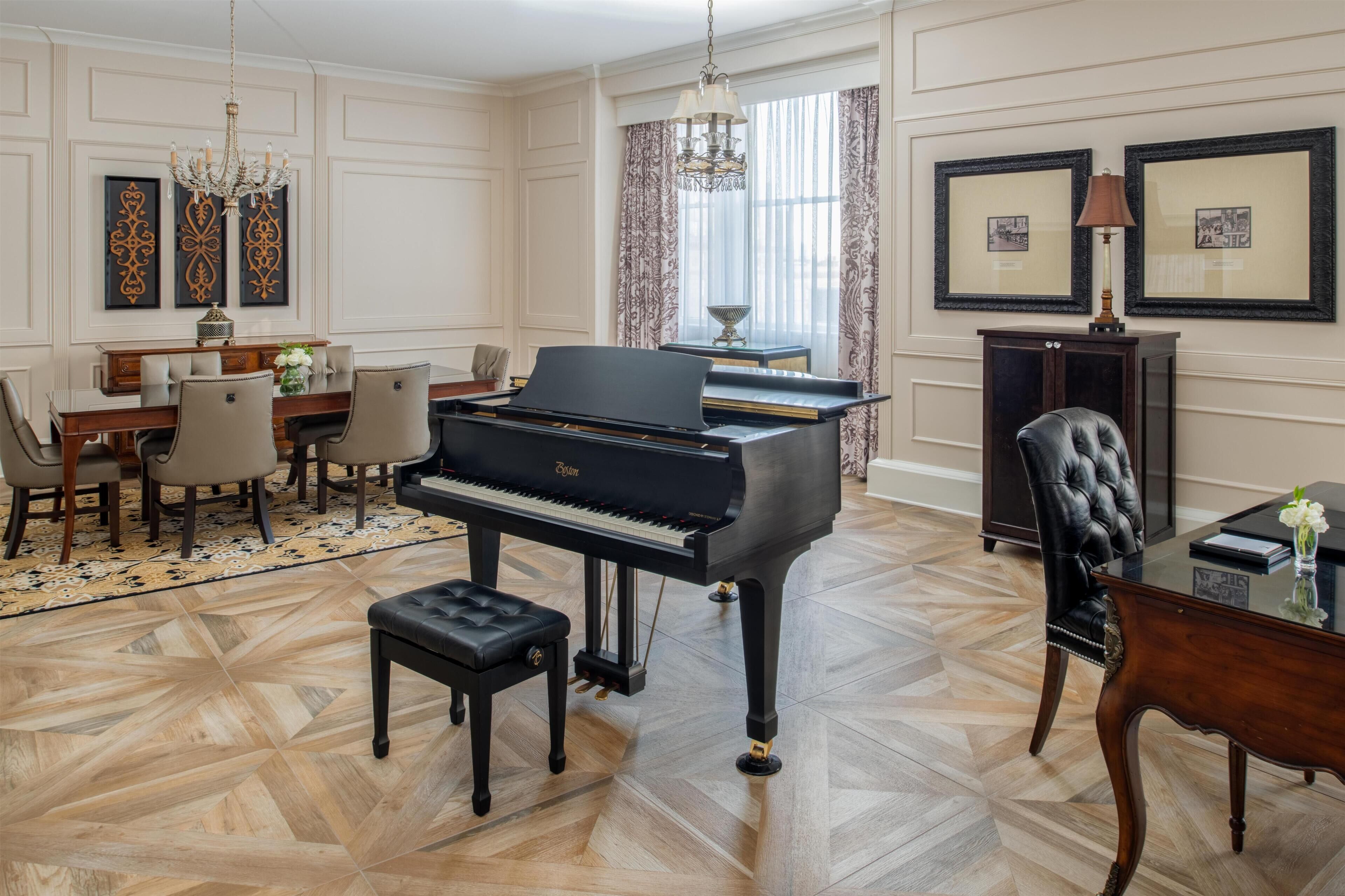 A grand piano in a well-appointed room with a dining area, chandelier, and framed pictures on the walls.