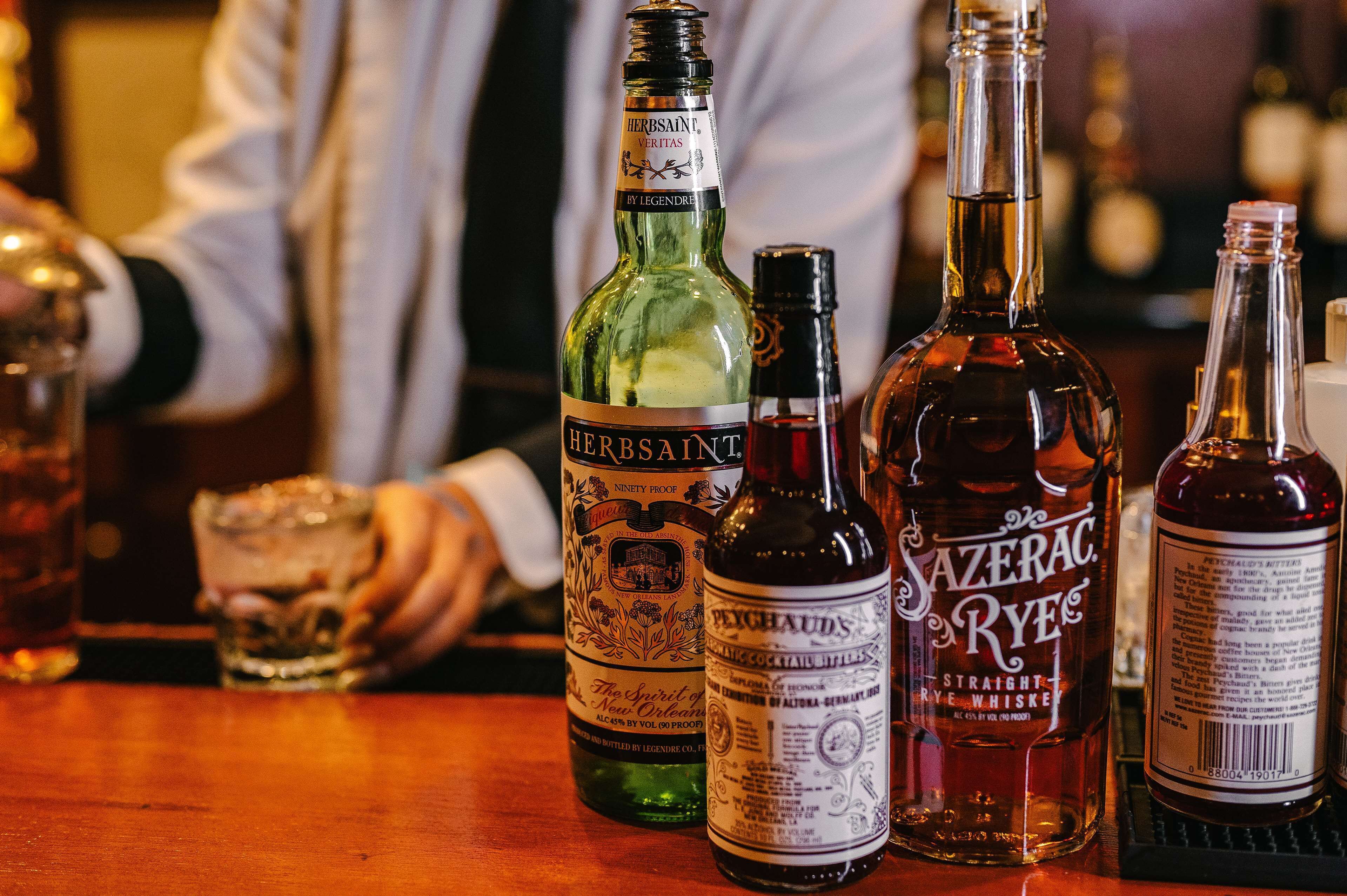 Eine Bar mit verschiedenen Spirituosenflaschen, darunter 'Herbsaint' und 'Sazerac Rye'.