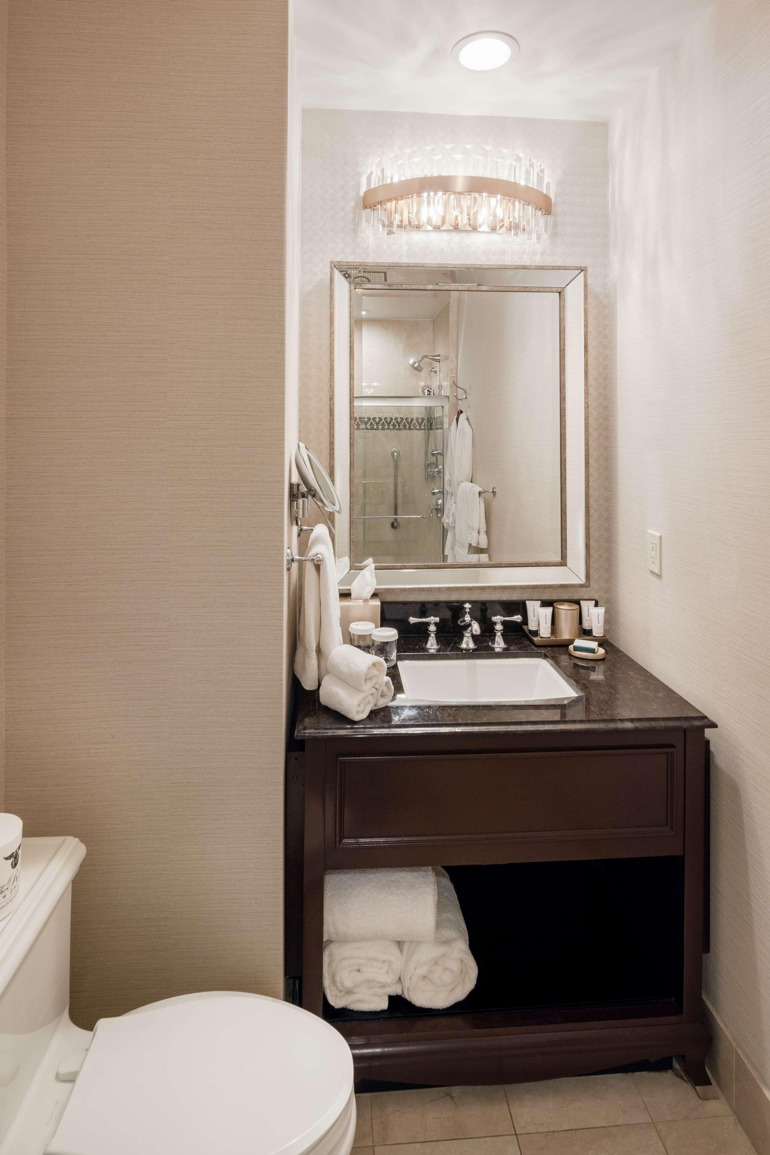 A bathroom with a white toilet, a dark brown vanity with a large mirror, a chandelier above the mirror, and a towel rack with folded towels.