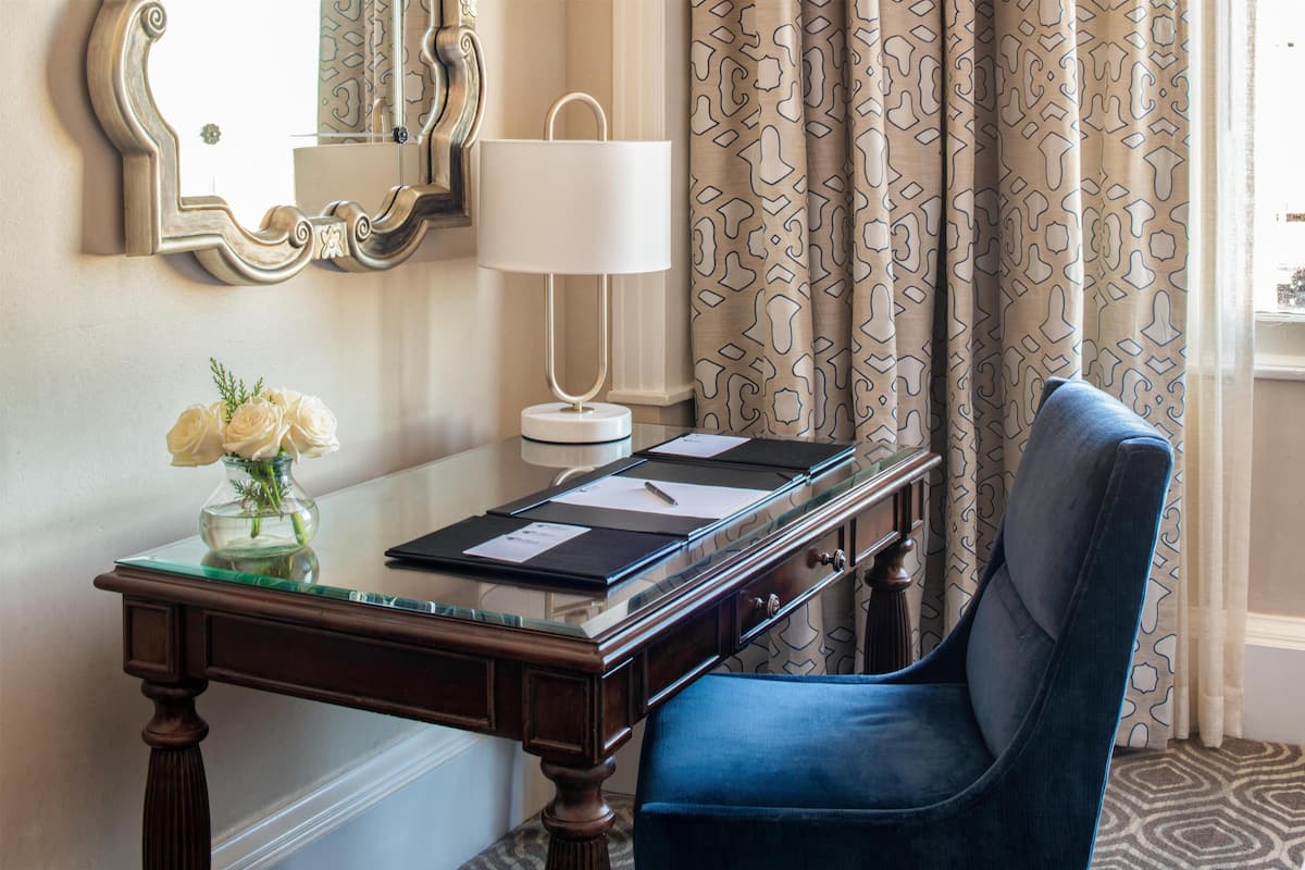 Een hotelkamer met een houten bureau, een blauwe stoel, een vaas met bloemen, een lamp en kamergordijnen met een patroon.