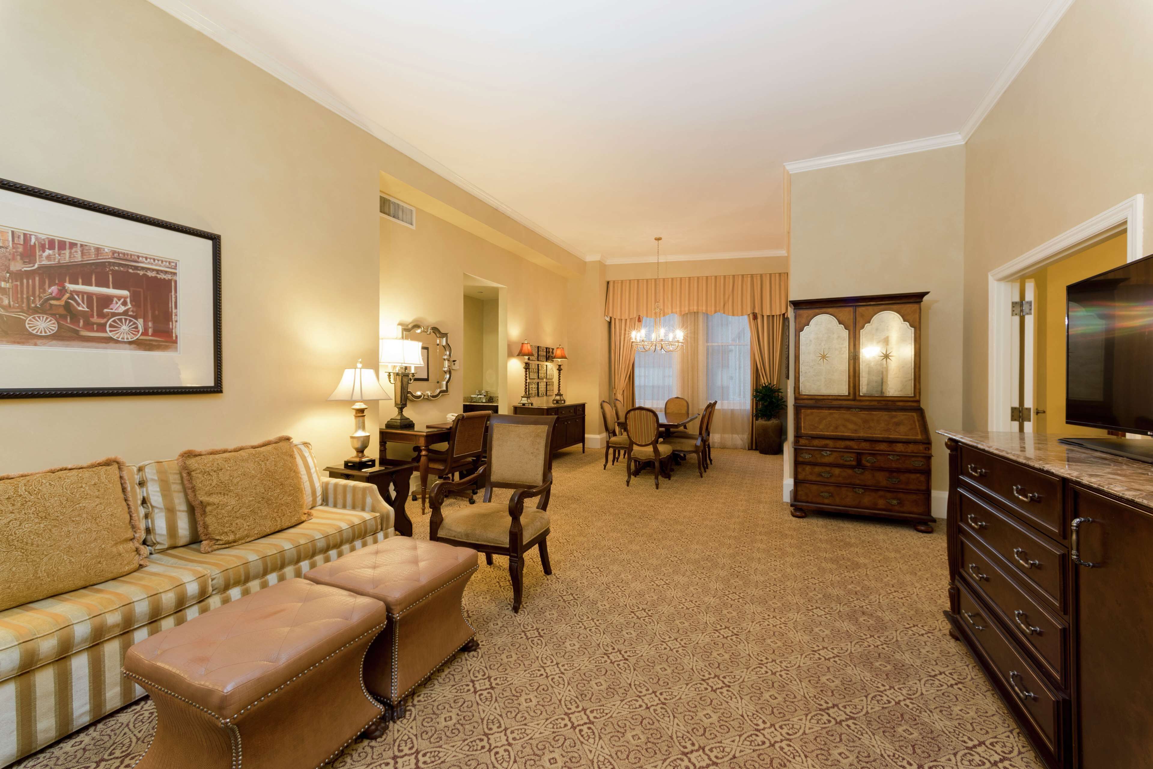 A hotel room with a sofa, ottoman, dining table, chairs, a dresser, and a television.