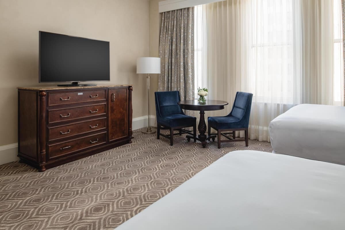 Hotelkamer met een televisie op een houten dressoir, twee stoelen rondom een kleine tafel, een bed en een raam met gordijnen.